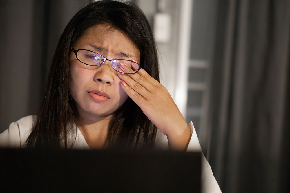 A woman wearing glasses rubs her eyes while working on a laptop in a dimly lit room, illustrating symptoms of eye strain or fatigue.