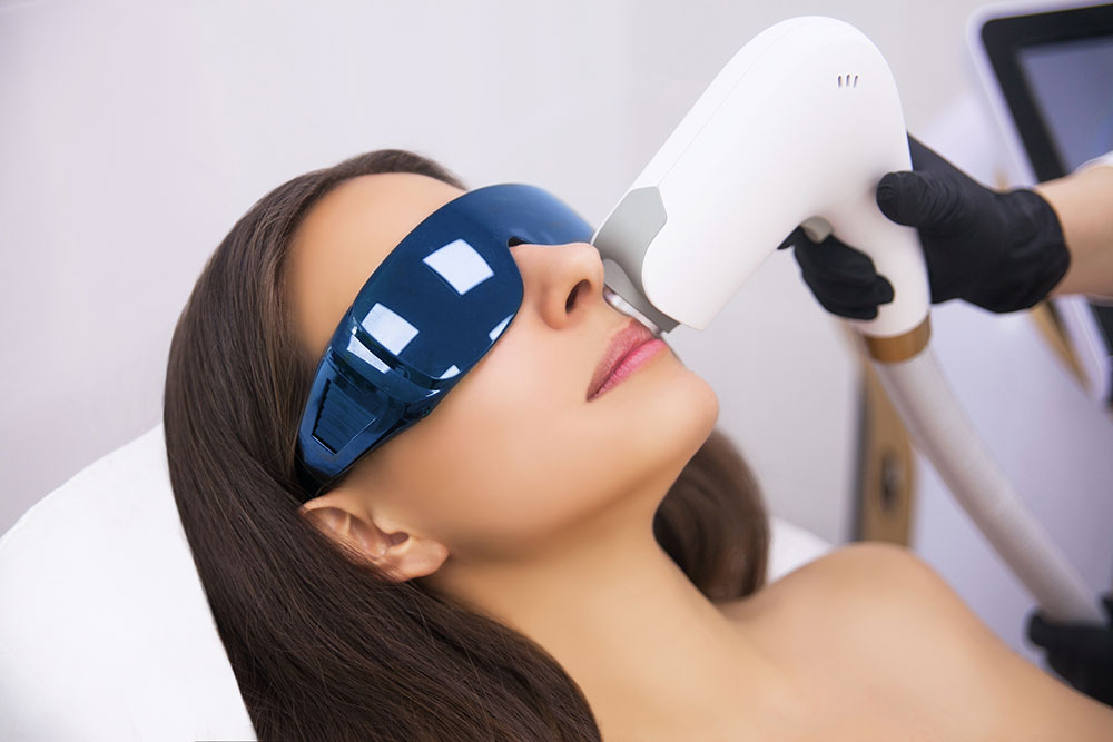 Woman receiving a skin treatment with a laser device while wearing protective eyewear. The setting indicates a clinical aesthetic procedure.