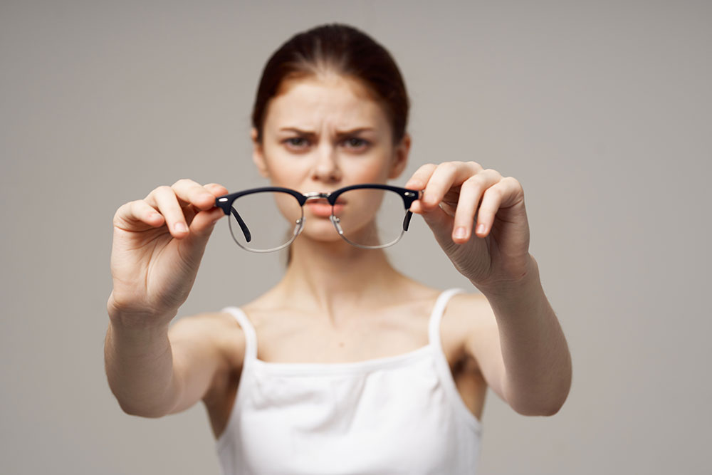 Woman holding glasses in front of her, looking frustrated. The image conveys themes of vision problems or dissatisfaction with eyewear.